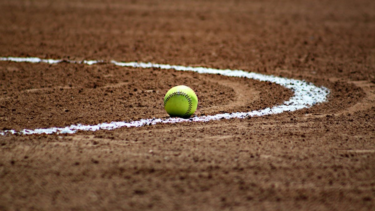 A yellow softball rests at the edge of a chalk line on a dirt infield