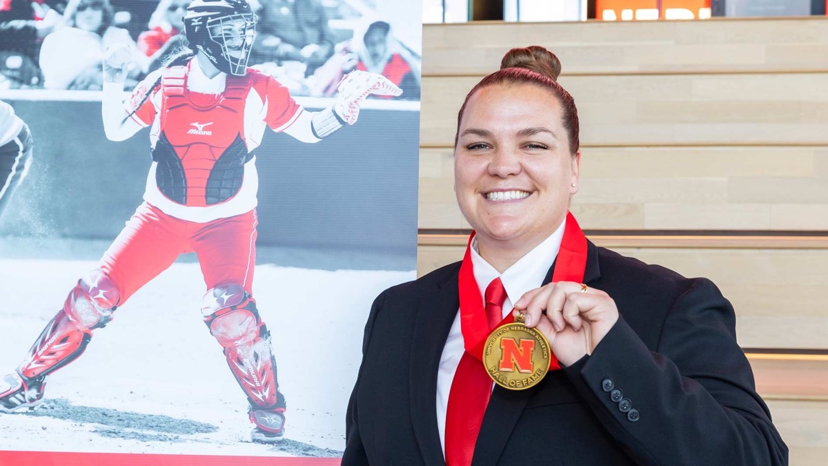 Taylor Edwards in a dark suit holds up her Nebraska Athletics Hall of Fame medal beside a photo of her catching for the Huskers